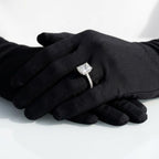 Radiant cut diamond engagement ring set in white gold or platinum with a hidden halo and pavé diamond band, displayed on black-gloved hands.