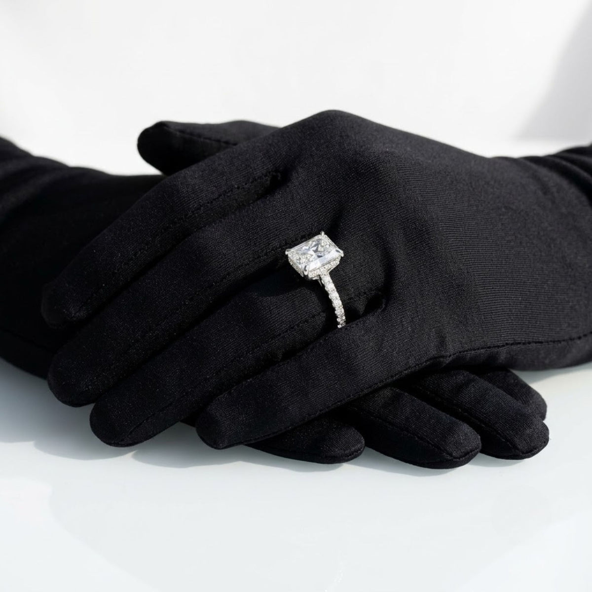Radiant cut diamond engagement ring set in white gold or platinum with a hidden halo and pavé diamond band, displayed on black-gloved hands.