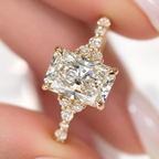 Close-up of fingers holding a yellow gold radiant cut diamond ring, showing the center stone, trio side clusters, and round diamonds along the band