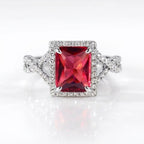 A silver-toned ring featuring a large rectangular red gemstone surrounded by a halo of small clear stones, with a split shank also accented by pavé stones, set against a clean white background.