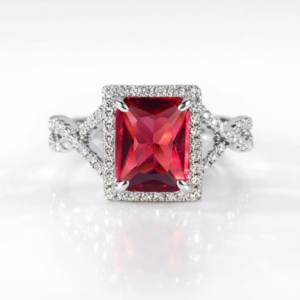 A silver-toned ring featuring a large rectangular red gemstone surrounded by a halo of small clear stones, with a split shank also accented by pavé stones, set against a clean white background.