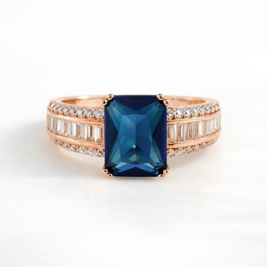 A rose gold ring featuring a large rectangular blue gemstone in the center, flanked by rows of small clear baguette and round-cut stones on a clean white background.