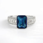 A silver-toned ring featuring a rectangular emerald-cut dark blue gemstone in the center, flanked by rows of small clear baguette and round-cut stones on a clean white background.