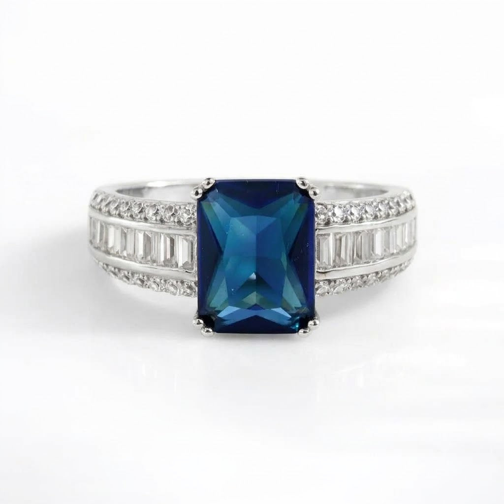 A silver-toned ring featuring a rectangular emerald-cut dark blue gemstone in the center, flanked by rows of small clear baguette and round-cut stones on a clean white background.
