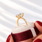 Side profile of a yellow gold four-prong radiant cut diamond engagement ring with baguette diamonds on the shank, resting in a burgundy velvet ring box held by red-manicured fingers