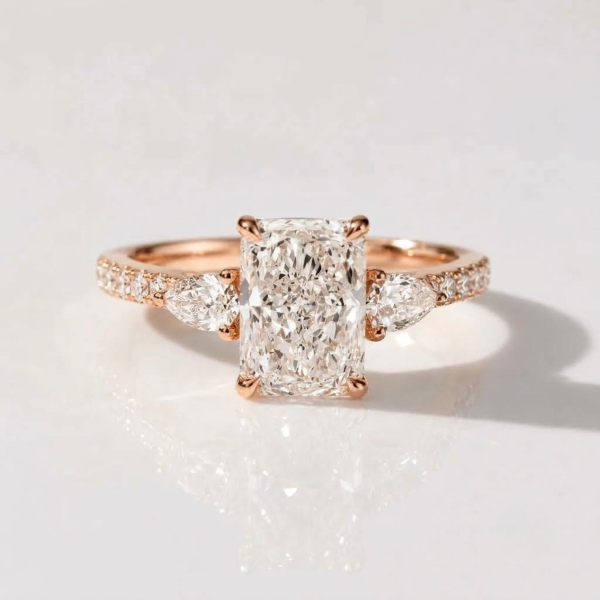 Ultra-realistic product photograph of an 18K rose gold three-stone engagement ring, showcasing a central radiant-cut diamond set between two smaller pear-cut diamonds. The ring features a diamond-pavé band and is displayed on a clean white background with subtle reflections.