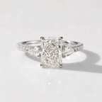 High-resolution product photograph of a platinum three-stone engagement ring, featuring a central radiant-cut diamond flanked by two pear-cut diamonds. The ring has a diamond-pavé band and is resting on a clean, reflective white surface with soft studio lighting.