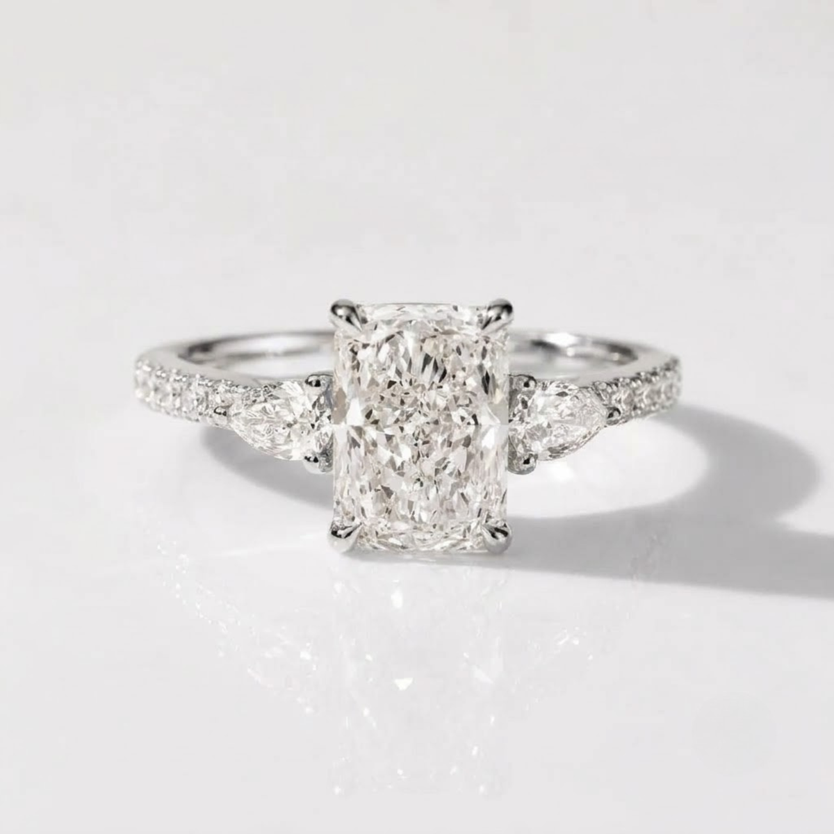 High-resolution product photograph of a platinum three-stone engagement ring, featuring a central radiant-cut diamond flanked by two pear-cut diamonds. The ring has a diamond-pavé band and is resting on a clean, reflective white surface with soft studio lighting.