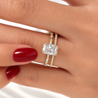 Hand with red nails wearing a yellow gold radiant cut diamond ring with baguette accents, stacked with a slim pavé diamond wedding band