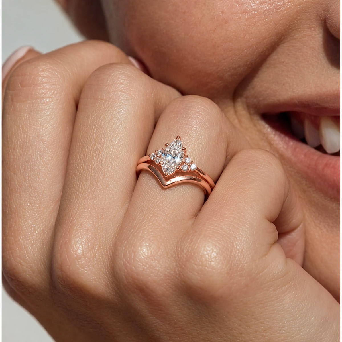 Close-up of a hand with a rose gold marquise-cut diamond cluster engagement ring stacked with a chevron wedding band, held near a smiling face