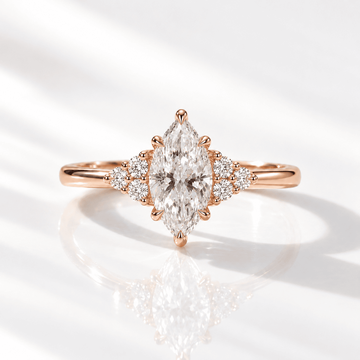Rose gold marquise-cut diamond ring with two trios of round side diamonds and a smooth polished band, resting on a reflective white surface in soft light.