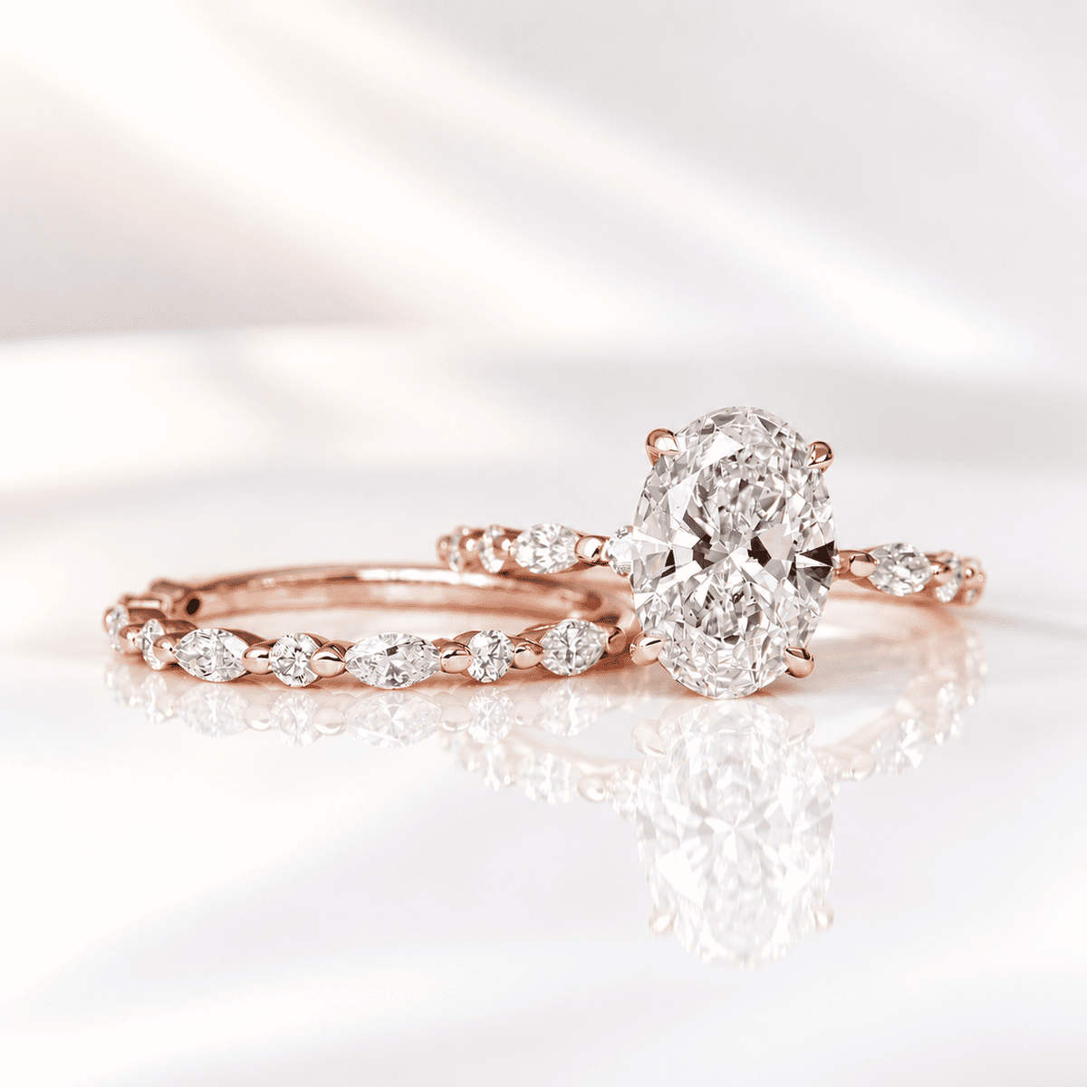 Rose gold oval diamond engagement ring stacked with a matching band of marquise and round accent diamonds, photographed on a reflective white surface in soft light.