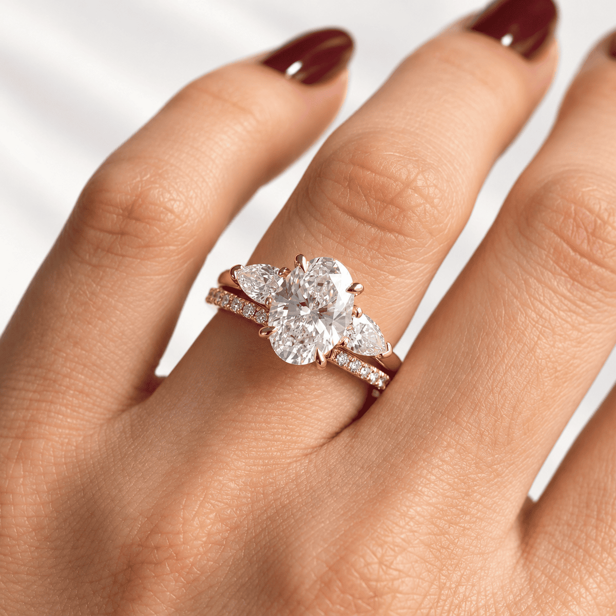 Hand with dark red nail polish wearing a rose gold ring with a center oval diamond, two pear-shaped side diamonds, and a pavé diamond band