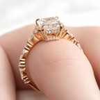 Close-up of a rose gold radiant cut diamond engagement ring on a finger, showing the pavé hidden halo beneath the center stone and marquise and round diamonds along the shank