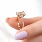 Side view of a rose gold radiant cut diamond engagement ring held between fingers with a lilac manicure, showing a hidden halo under the center stone, a trio of round diamonds, and alternating marquise and round diamonds along the shank