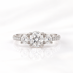 Round cut diamond center stone in a four-prong white gold setting, flanked by a round side stone on each side with a pavé diamond band
