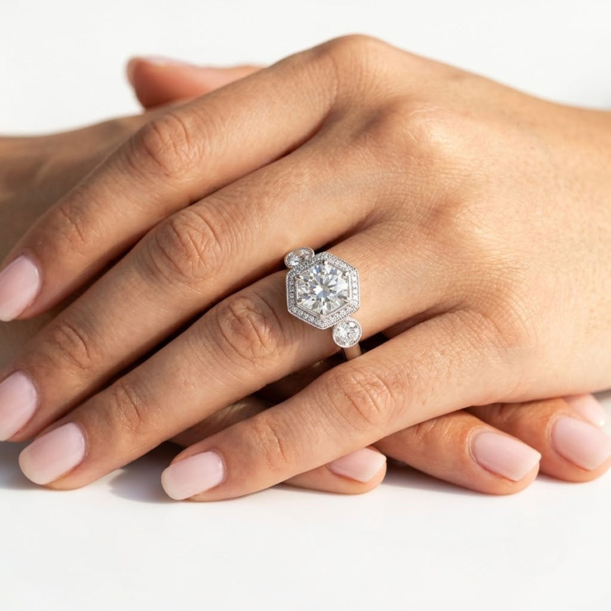 Vintage-inspired round cut diamond engagement ring set in white gold, featuring a hexagonal halo with milgrain detailing and round bezel-set side accents, worn on a hand.