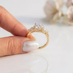 Three-stone yellow gold engagement ring held between fingers, featuring a round brilliant cut center stone flanked by two pear cut side diamonds in an open trellis setting.