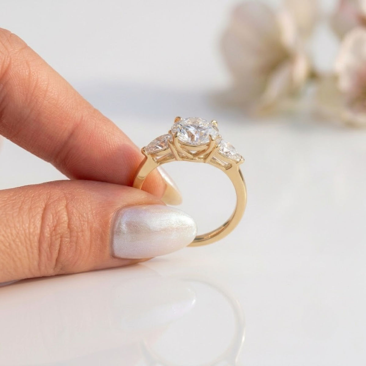 Three-stone yellow gold engagement ring held between fingers, featuring a round brilliant cut center stone flanked by two pear cut side diamonds in an open trellis setting.