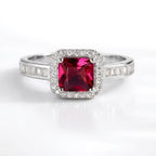 A silver-toned ring featuring a square-cut deep red center stone surrounded by a halo of small clear stones, with additional clear stones set along the band, shown on a clean white background.