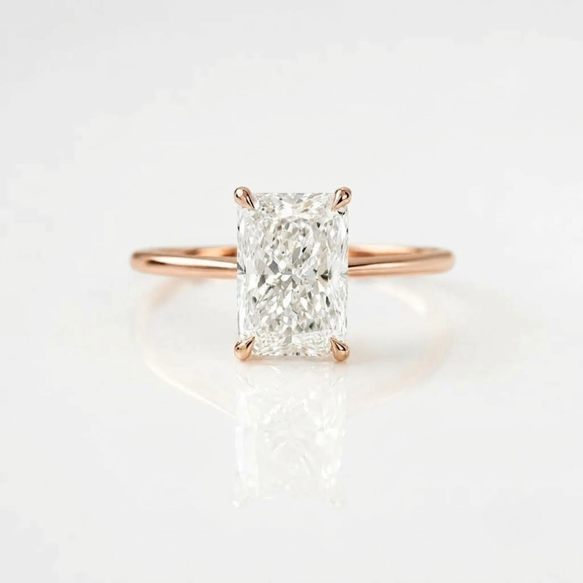 High-quality, ultra-realistic product photograph of a solitaire engagement ring featuring a large, radiant-cut center diamond held by four prongs. The ring has a simple, polished 18K rose gold vermeil band with a luxurious warm pink-gold tone. It is positioned in a balanced, lying position on a clean pure white background with soft studio lighting and subtle natural shadows. The reflection of the ring and diamond is visible on the surface.