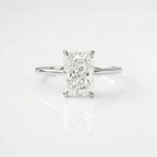 High-quality, ultra-realistic product photograph of a solitaire engagement ring featuring a large, radiant-cut center diamond held by four prongs. The ring has a simple, polished silver band. It is positioned in a balanced, lying position on a clean pure white background with soft studio lighting and subtle natural shadows. The reflection of the ring and diamond is visible on the surface.