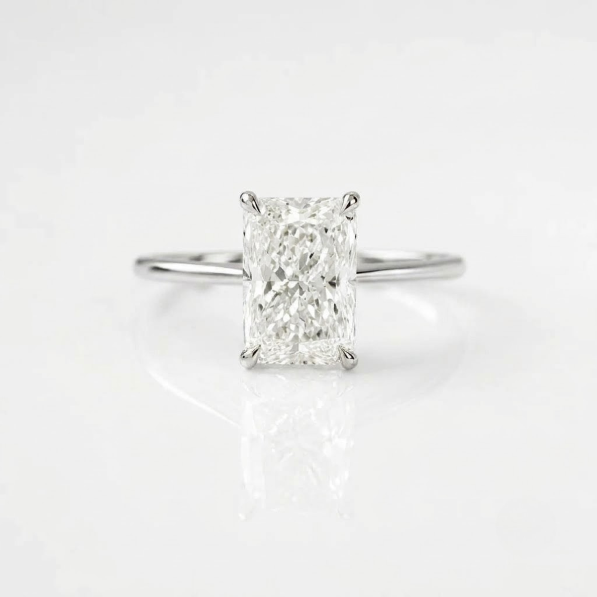 High-quality, ultra-realistic product photograph of a solitaire engagement ring featuring a large, radiant-cut center diamond held by four prongs. The ring has a simple, polished silver band. It is positioned in a balanced, lying position on a clean pure white background with soft studio lighting and subtle natural shadows. The reflection of the ring and diamond is visible on the surface.