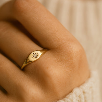 A close-up view of a gold signet ring featuring a delicate engraved starburst design with a sparkling round diamond set in the center,
