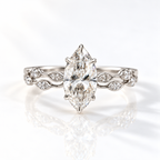 White gold marquise diamond engagement ring with six prongs, paired with a wedding band of bezel-set marquise and round diamonds, shown on a soft reflective surface