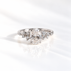 White gold oval diamond engagement ring with scattered round diamond clusters along the shoulders of a slim band, shown on a soft reflective surface