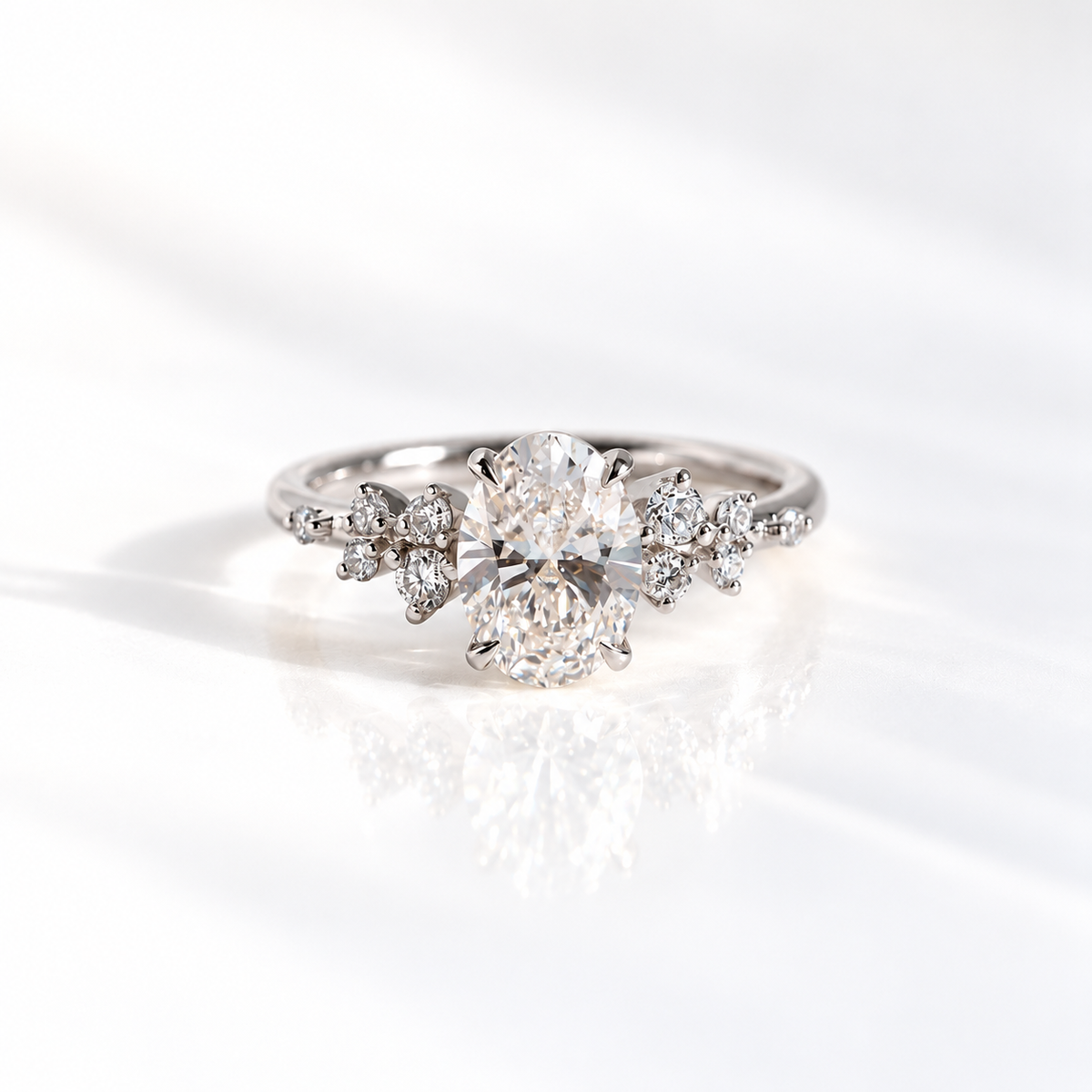 White gold oval diamond engagement ring with scattered round diamond clusters along the shoulders of a slim band, shown on a soft reflective surface