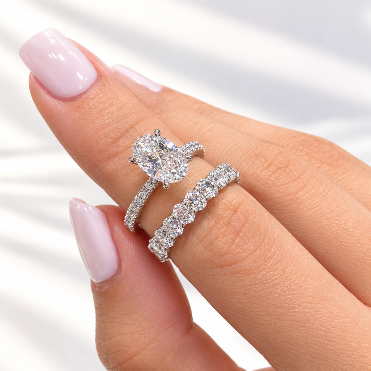 Hand with a light pink manicure wearing a white gold oval diamond engagement ring with a pavé shank, stacked with a wedding band of oval and round diamonds