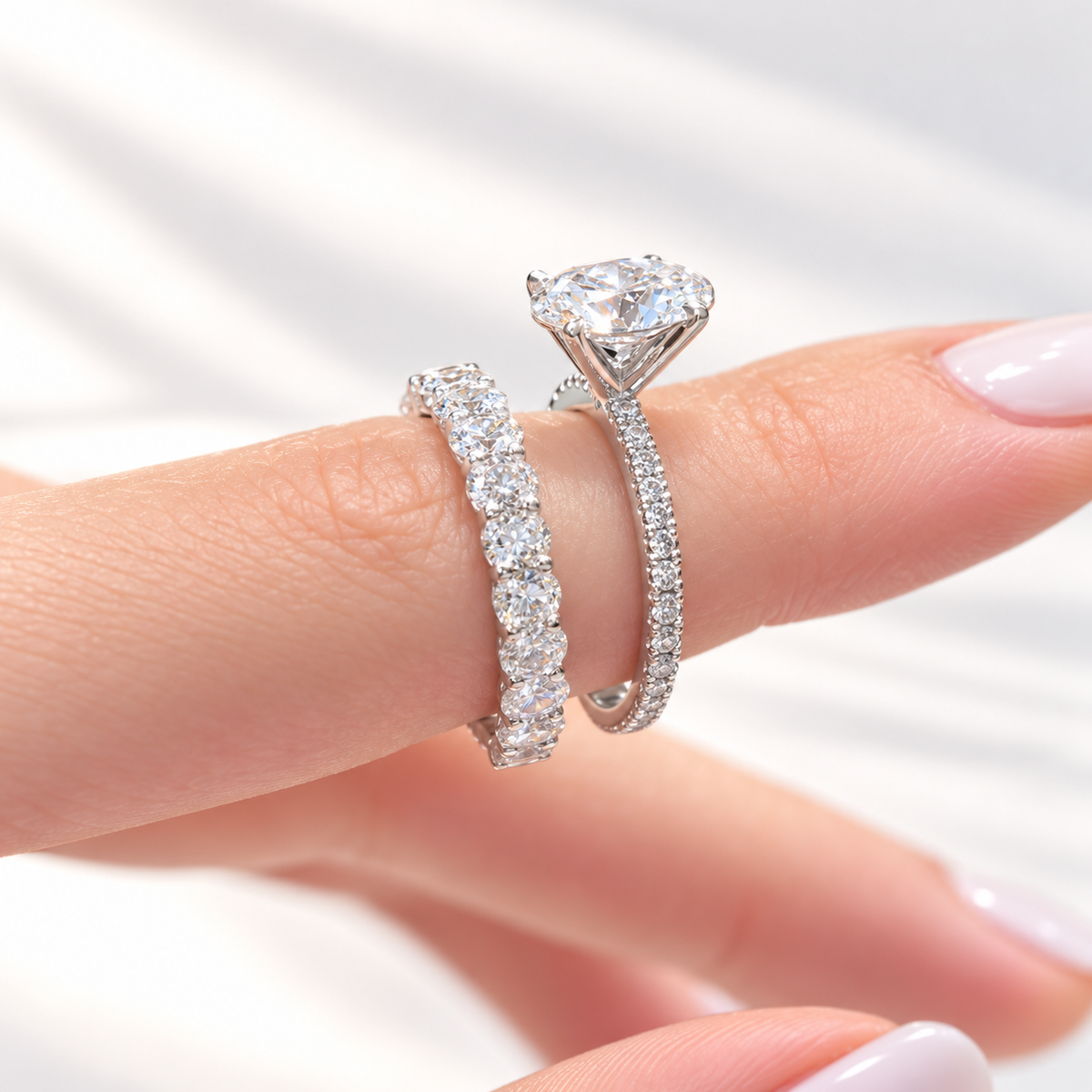 Fingers with a French manicure holding a white gold oval diamond engagement ring with a pavé shank and a matching wedding band of oval diamonds