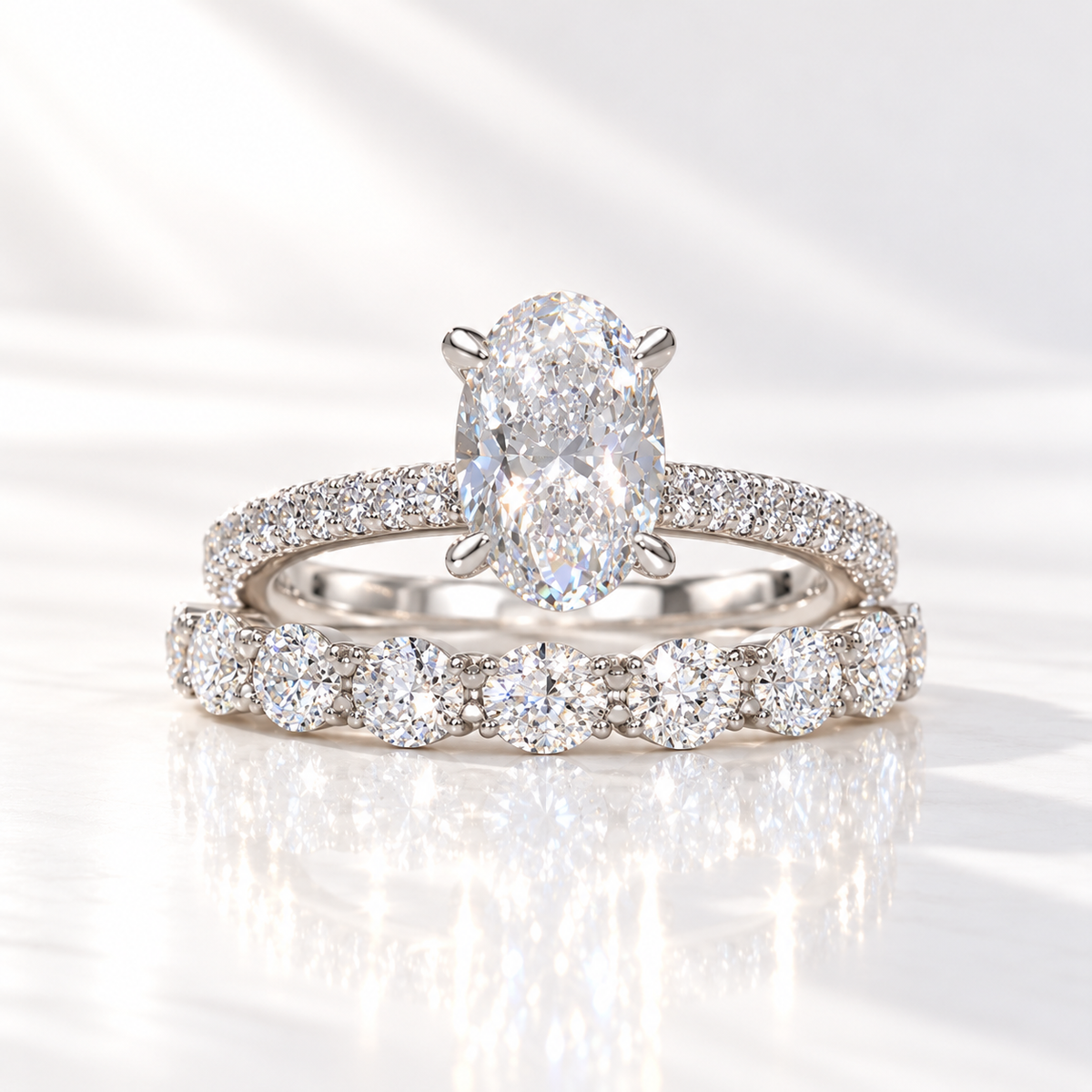 White gold oval diamond engagement ring with four prongs and a pavé diamond shank, stacked with a matching wedding band of large round diamonds, on a glossy white surface