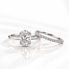 White gold oval diamond solitaire ring with a double prong setting, beside a matching pavé diamond wedding band, shown on a soft reflective surface