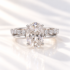 White gold oval diamond engagement ring with a four-prong setting, paired with a wedding band of round diamonds, shown on a soft reflective surface