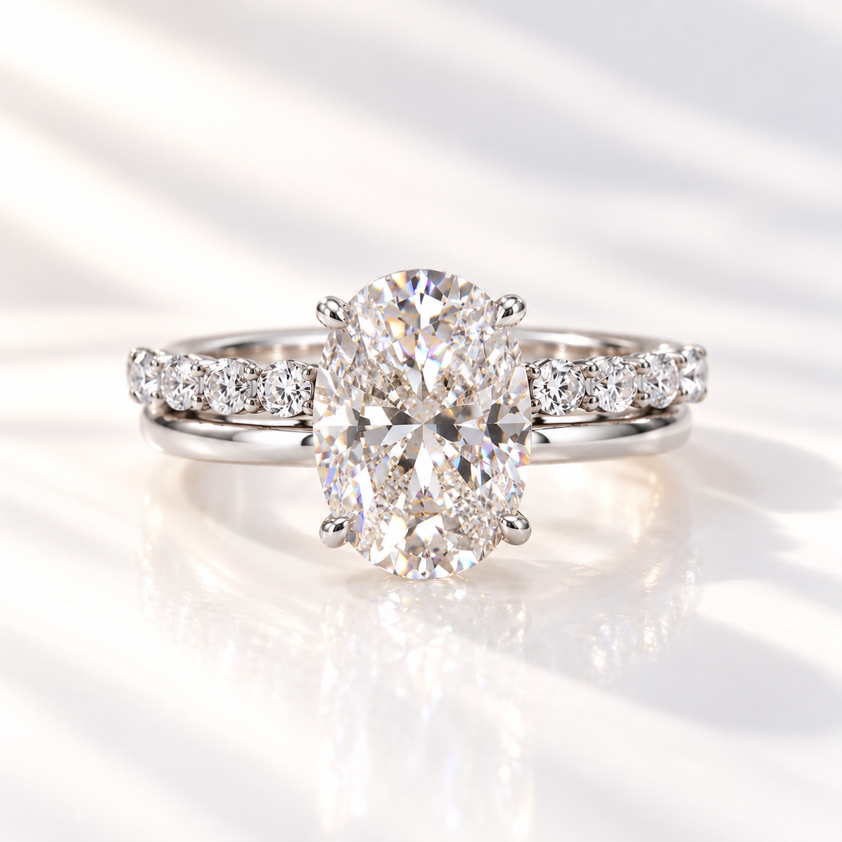 White gold oval diamond engagement ring with a four-prong setting, paired with a wedding band of round diamonds, shown on a soft reflective surface