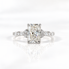 White gold radiant cut diamond engagement ring with four prongs, small round diamond trios on either side, and a delicate diamond-accented band