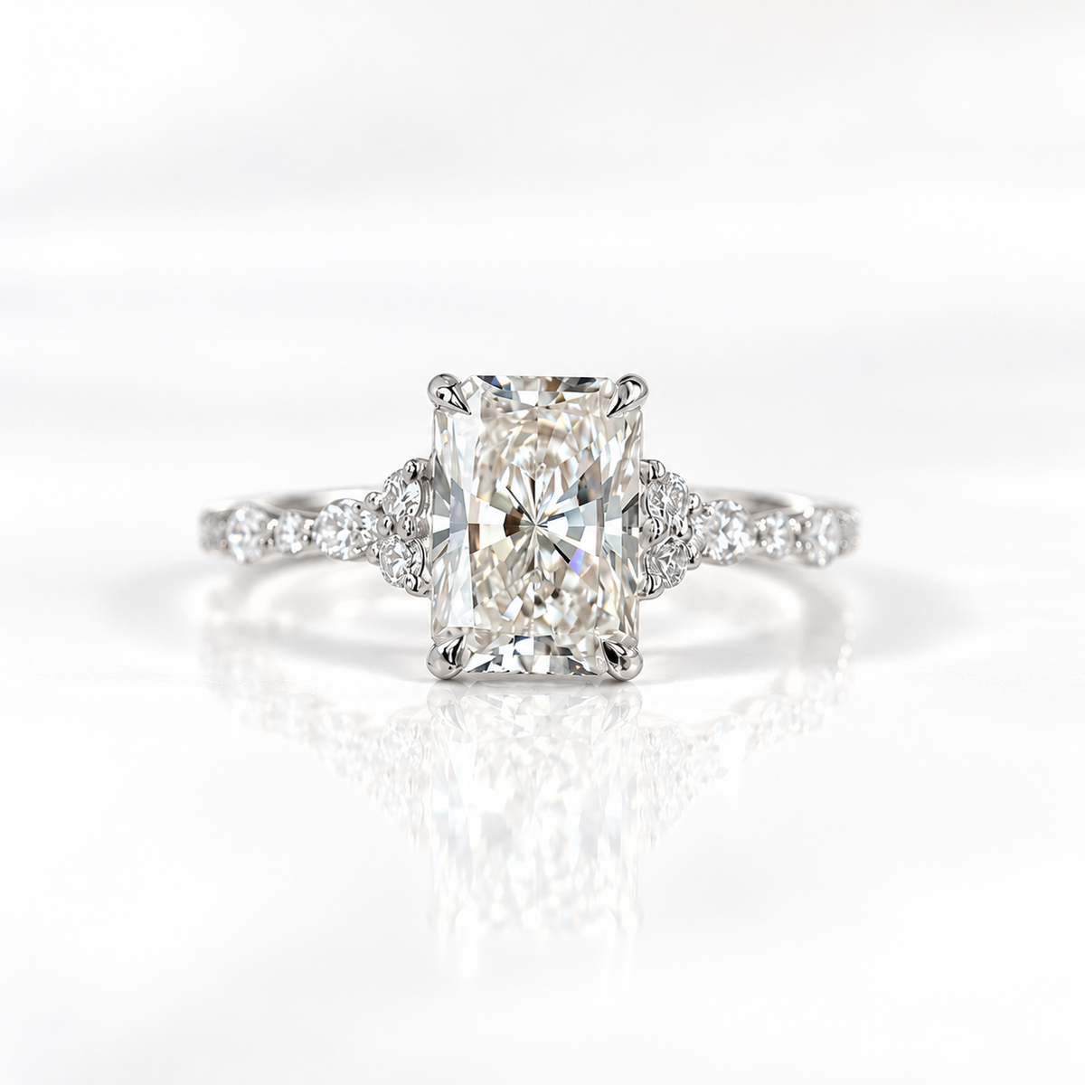 White gold radiant cut diamond engagement ring with four prongs, small round diamond trios on either side, and a delicate diamond-accented band