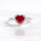 A heart-shaped red gemstone ring set in a silver-colored halo with small clear stones, shown on a clean white reflective surface.