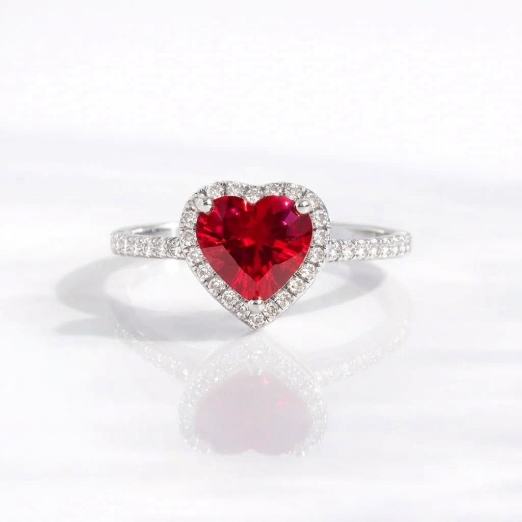 A heart-shaped red gemstone ring set in a silver-colored halo with small clear stones, shown on a clean white reflective surface.