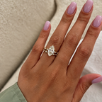 Hand with lilac nails wearing a yellow gold marquise diamond engagement ring and a matching wedding band of bezel-set marquise and round diamonds, against a cream upholstered background