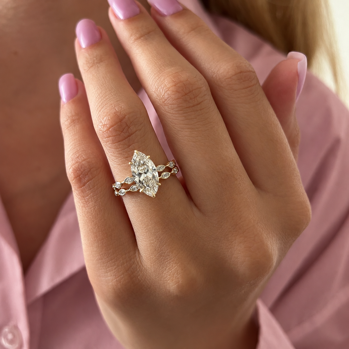 Hand with lilac nails wearing a yellow gold marquise diamond engagement ring with a stacked wedding band of bezel-set marquise and round diamonds, in front of a pink shirt