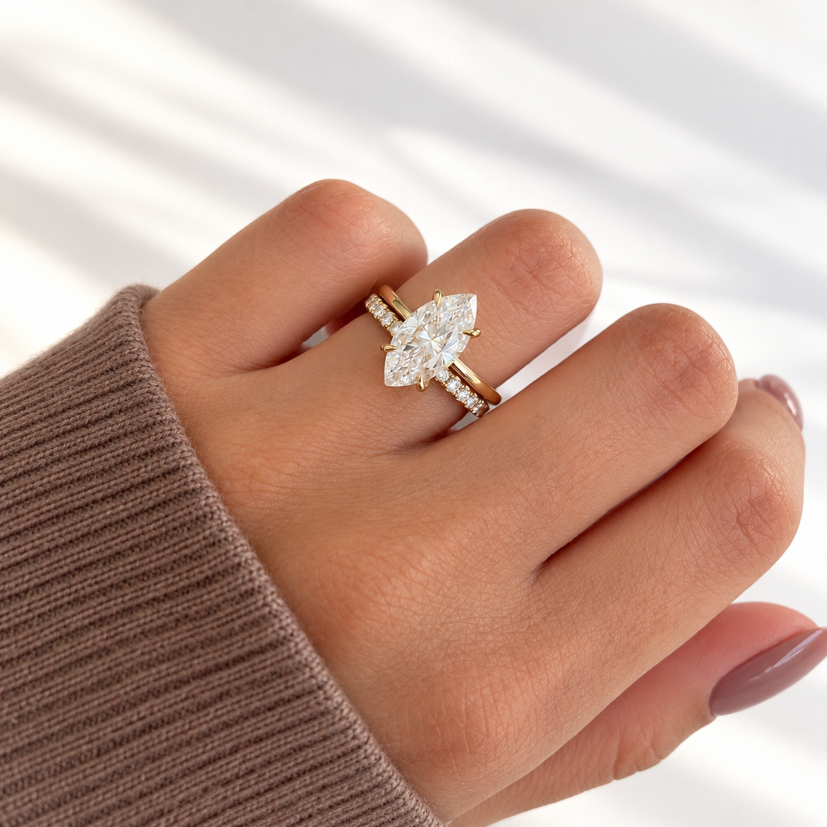 Hand in a brown knit sleeve wearing a yellow gold marquise diamond solitaire engagement ring with a six-prong setting, stacked with a slim pavé diamond wedding band