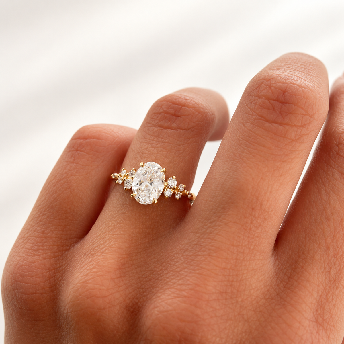 Hand wearing a yellow gold oval diamond engagement ring with symmetrical clusters of small round diamonds on either side of the center stone