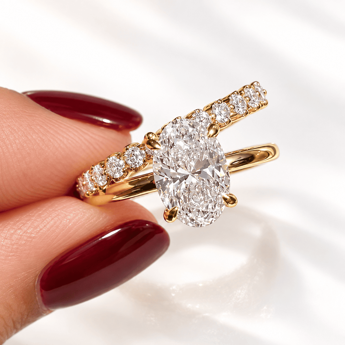 A hand with deep red manicured nails holding a yellow gold bypass ring, with an oval center diamond and a row of graduated round diamonds catching the light.