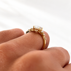 Angled side view of a yellow gold oval diamond ring on a finger, showing the four-prong setting and scattered round diamond accents on the band