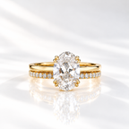Front view of a yellow gold oval diamond solitaire engagement ring with double prongs, stacked on a pavé diamond wedding band, on a glossy white surface