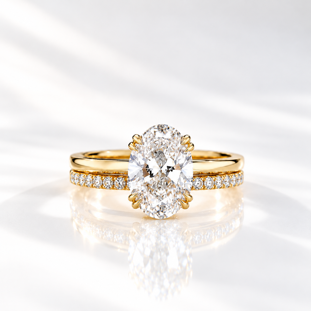 Front view of a yellow gold oval diamond solitaire engagement ring with double prongs, stacked on a pavé diamond wedding band, on a glossy white surface