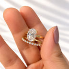 Close-up of fingers holding a yellow gold four-prong oval diamond engagement ring stacked with a delicate round diamond wedding band